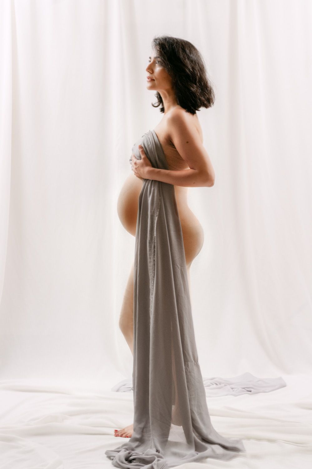 Pregnant woman draped in soft gray fabric posing elegantly in a bright white studio for a maternity photoshoot.