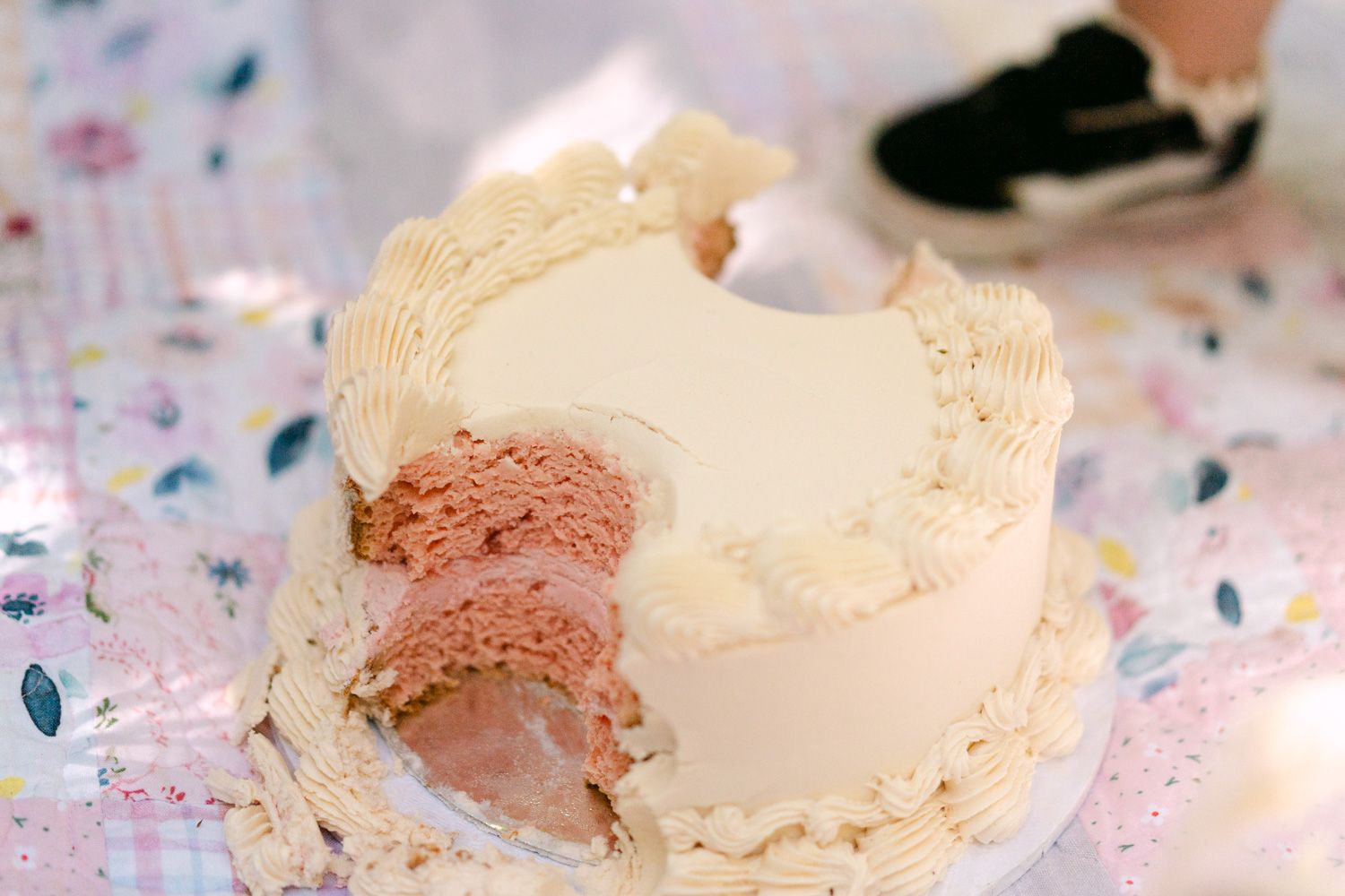 A white frosted layer cake with pink interior, partially cut open, displayed on a floral tablecloth.
