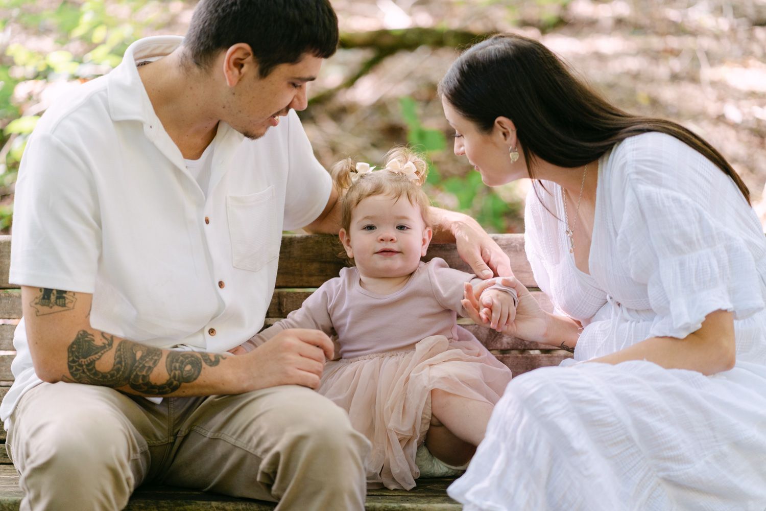 A young family with tattooed father, mother in white dress, and baby girl sitting on a wooden bench outdoors.
