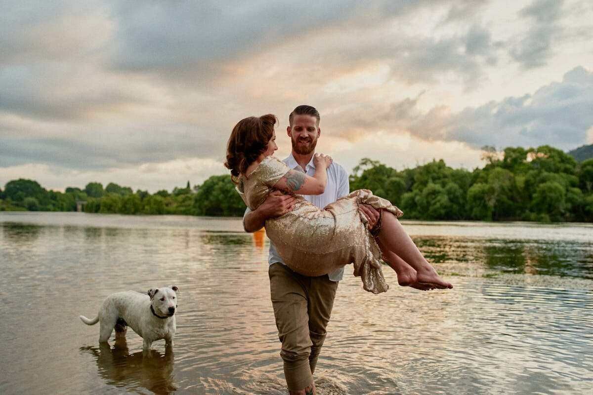 Man carrying woman in his arms while standing in a lake at sunset, with a white dog beside them.