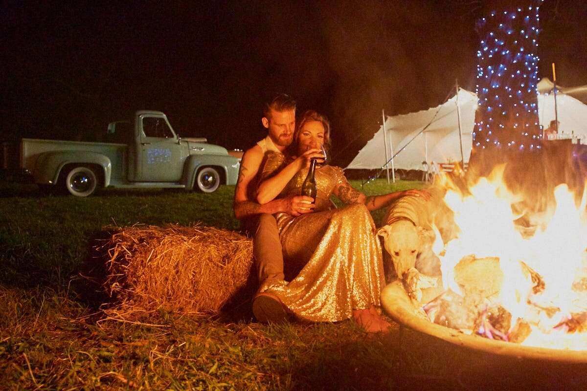 Couple sitting on hay bale by campfire at night, woman in gold dress, vintage truck and lit tent in background.