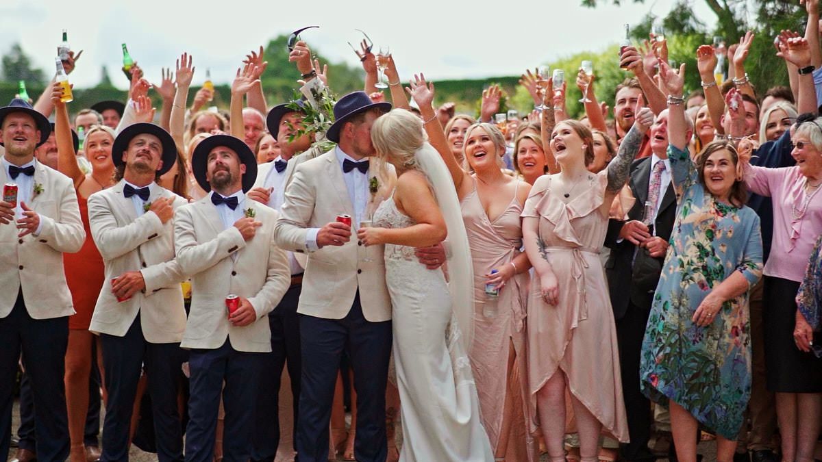 Joyful wedding celebration with bride and groom kissing surrounded by cheering guests in elegant attire outdoors.