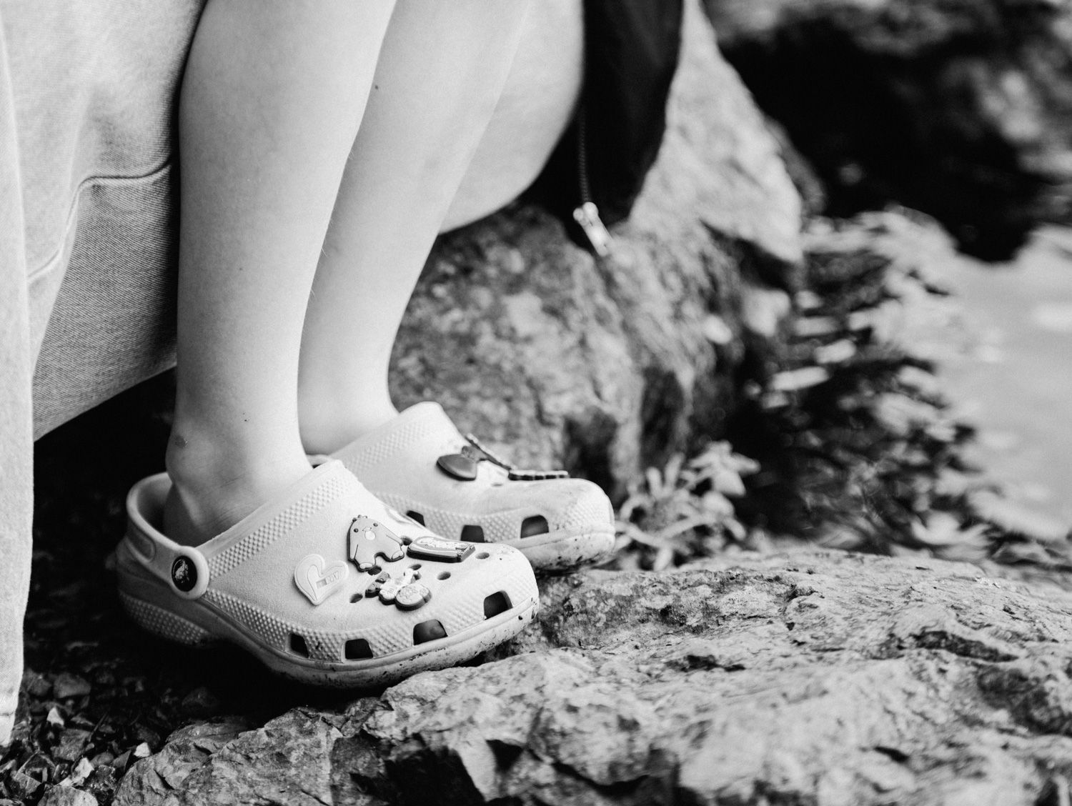 Close-up of child's feet wearing white Crocs sandals with charms, standing on rocky surface outdoors.