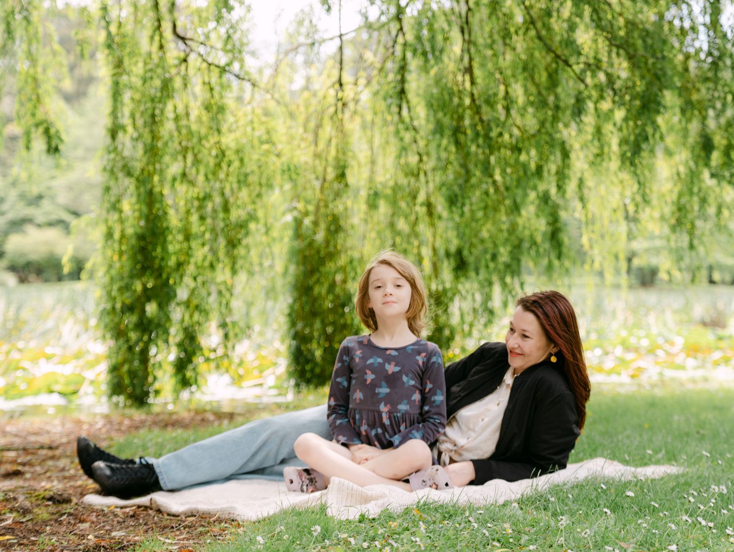 Two women sitting on a blanket under a weeping willow tree in a lush green park setting.