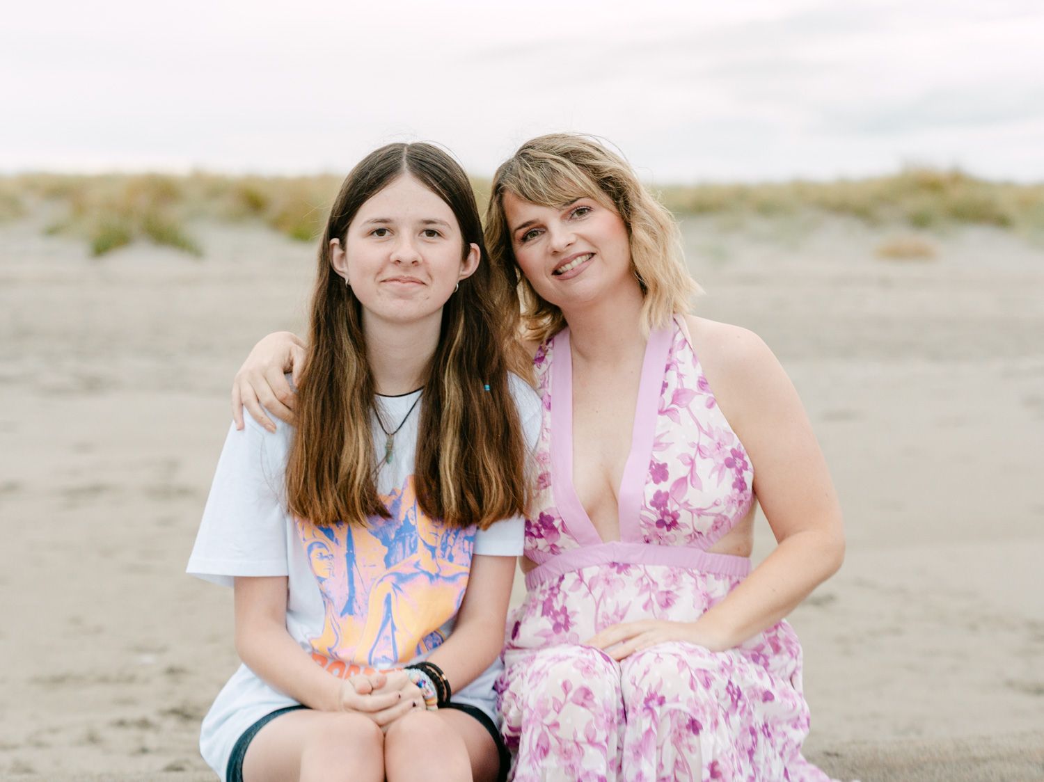 Two women smiling together on a sandy beach, one in a floral dress and one in a white tee.