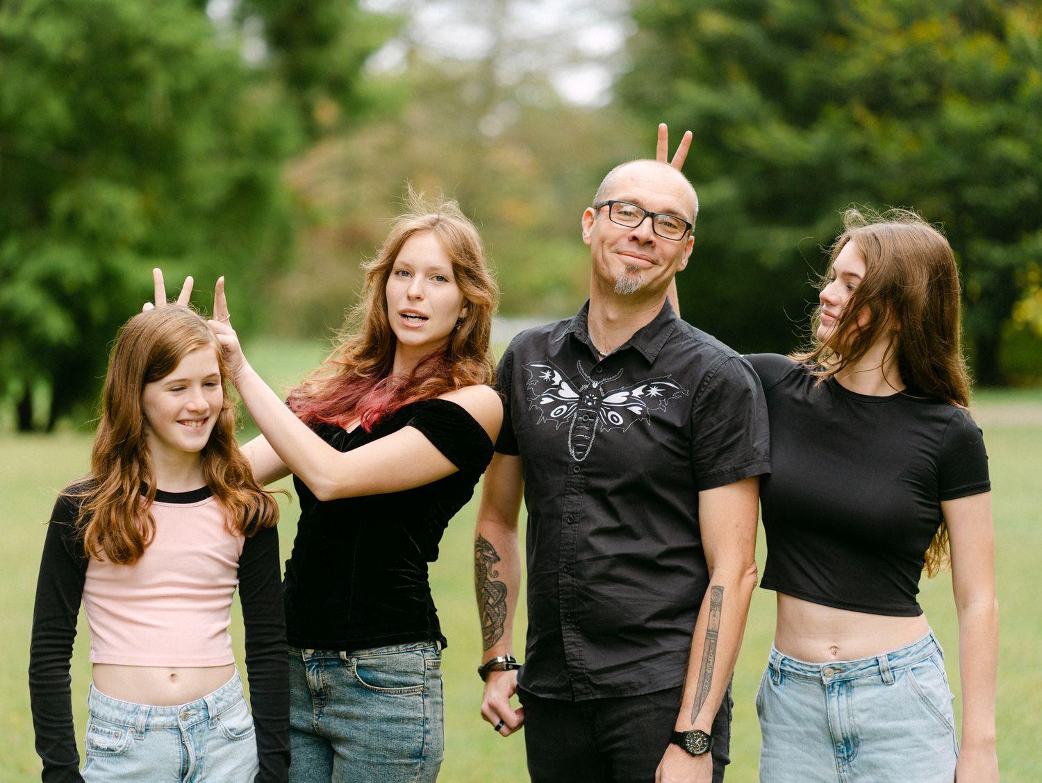 A man and three girls pose outdoors in a park, playfully making bunny ears behind each other's heads.
