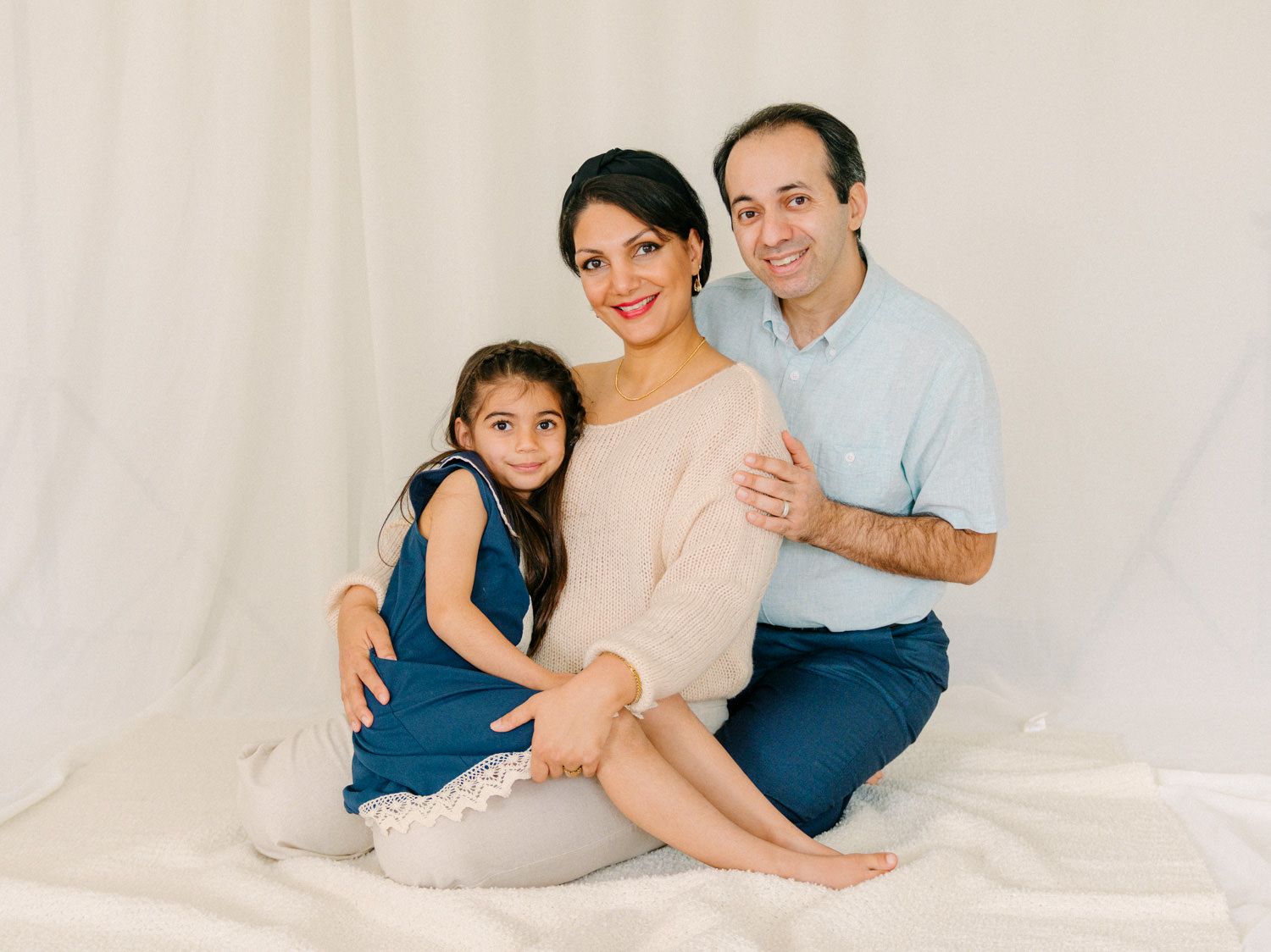 A smiling family of three poses together on a white backdrop, featuring a couple and their young daughter in a blue dress.