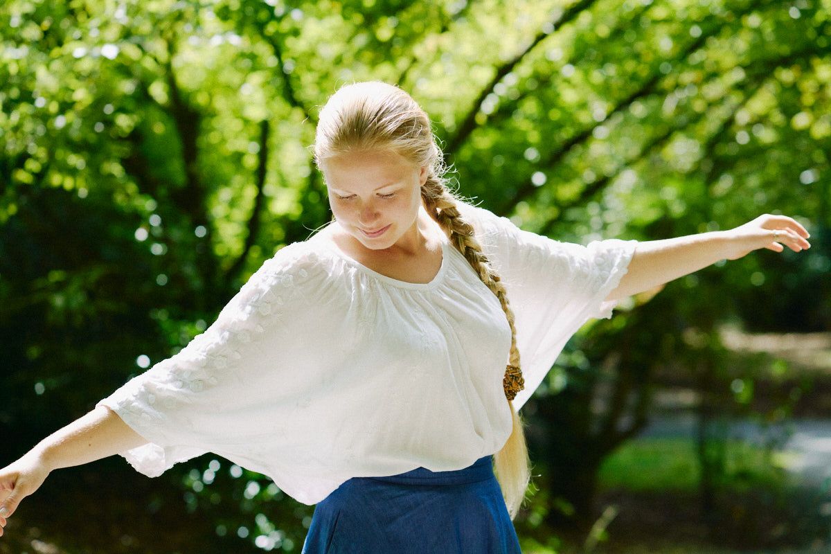 Young woman with braided hair in white blouse and blue skirt dancing outdoors in a lush green park.