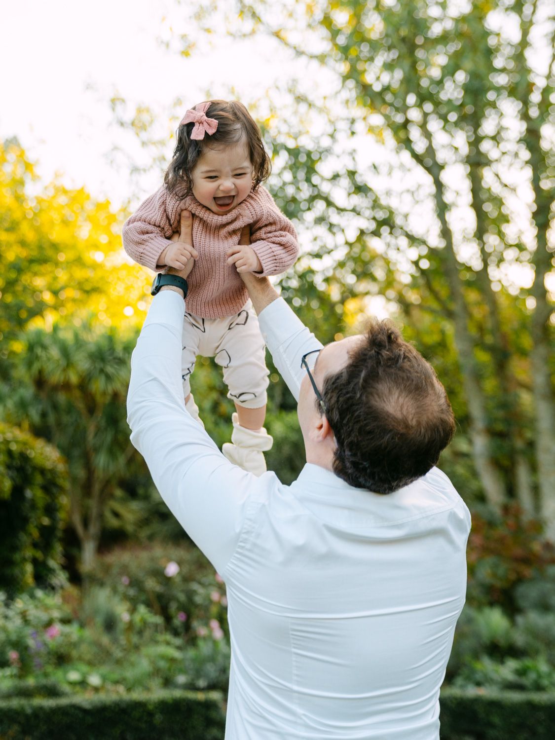 A father in white lifts his laughing toddler girl high in a lush green garden during a joyful outdoor family moment.