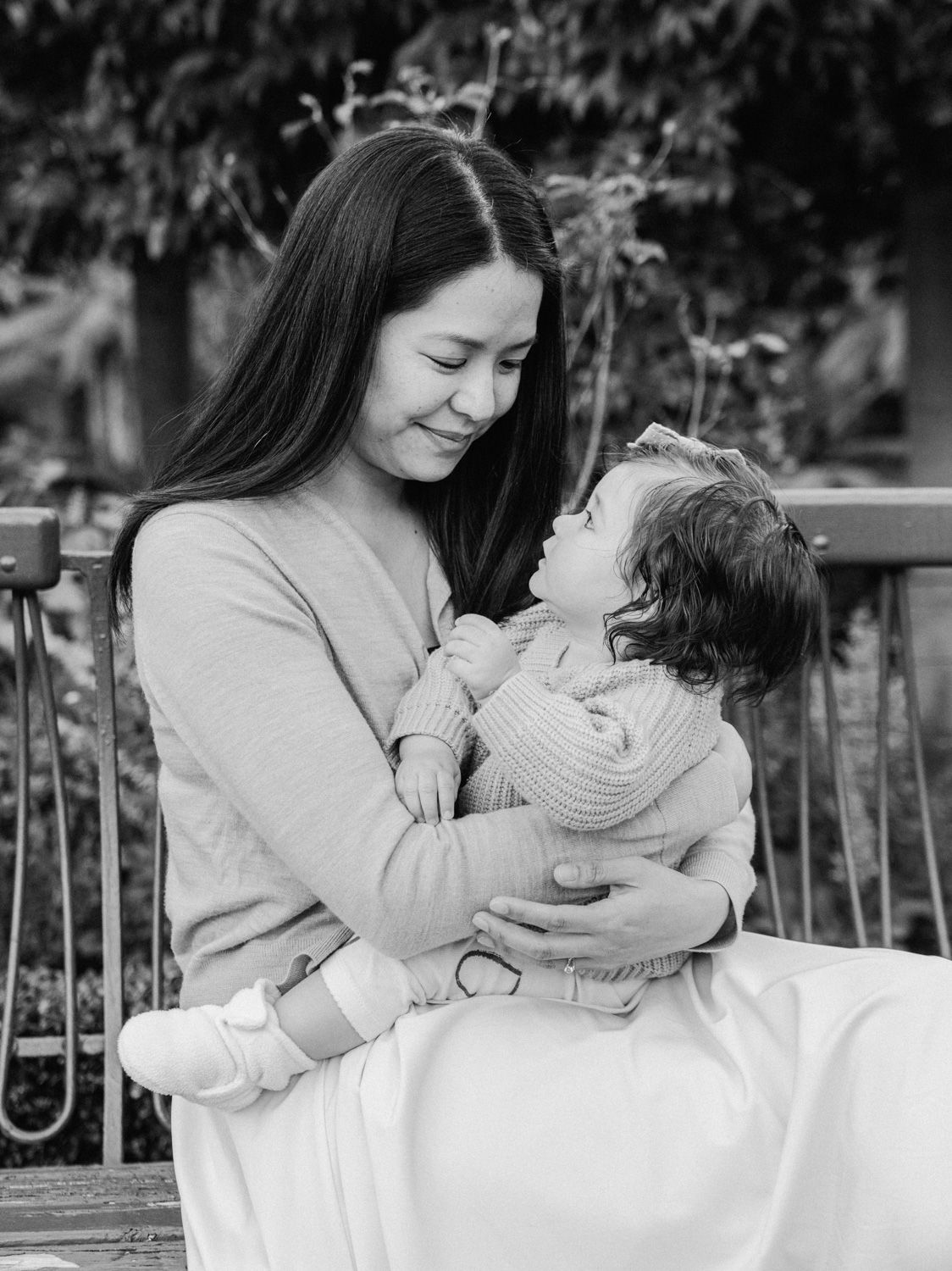 Black and white photo of a smiling woman holding a happy toddler on her lap outdoors on a bench.