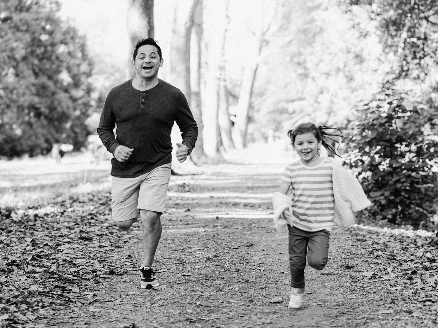 A man and young child joyfully running together on a tree-lined dirt path in a black and white photo.