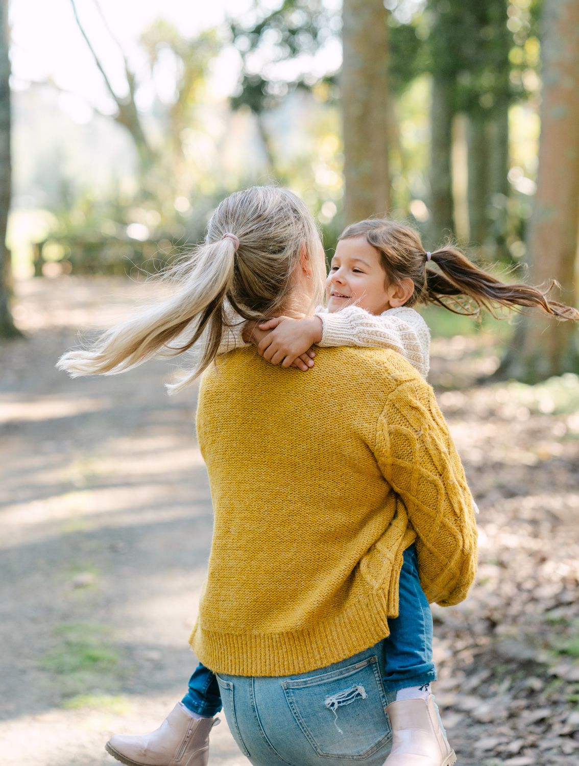 A mother in a yellow sweater holds her smiling daughter in a sunlit forest path, both with flowing hair.