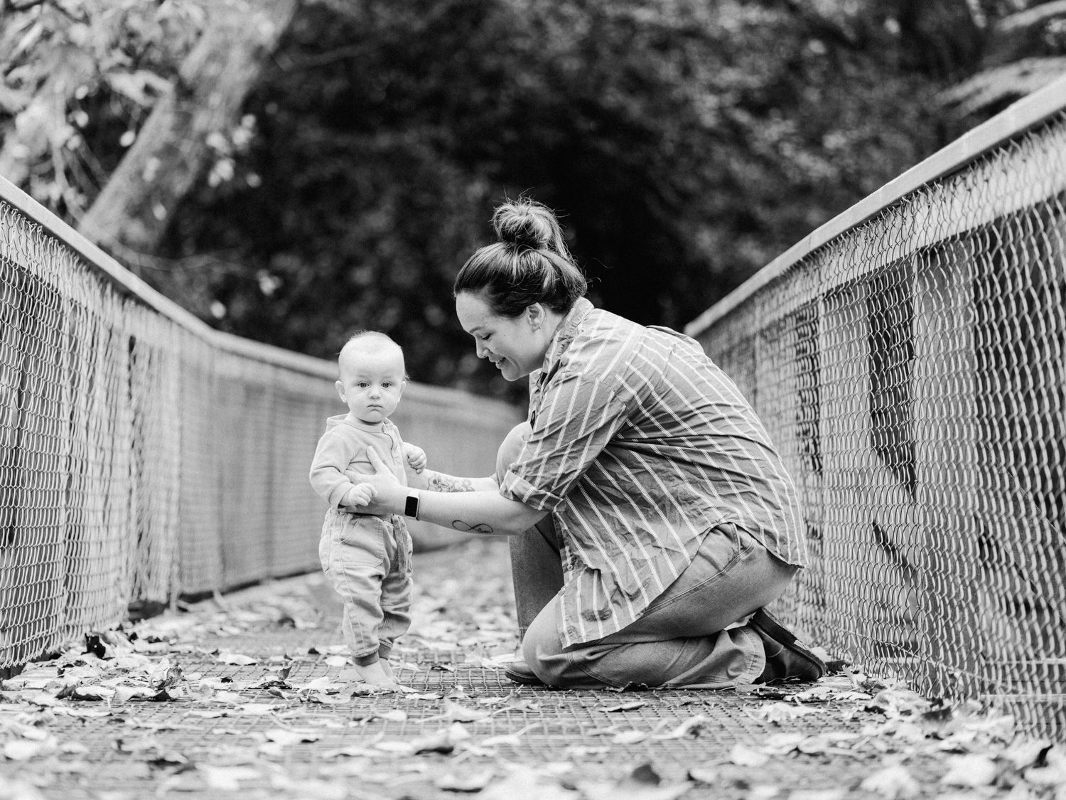 Black and white photo of a mother kneeling on a bridge, gently holding her toddler's hands outdoors.