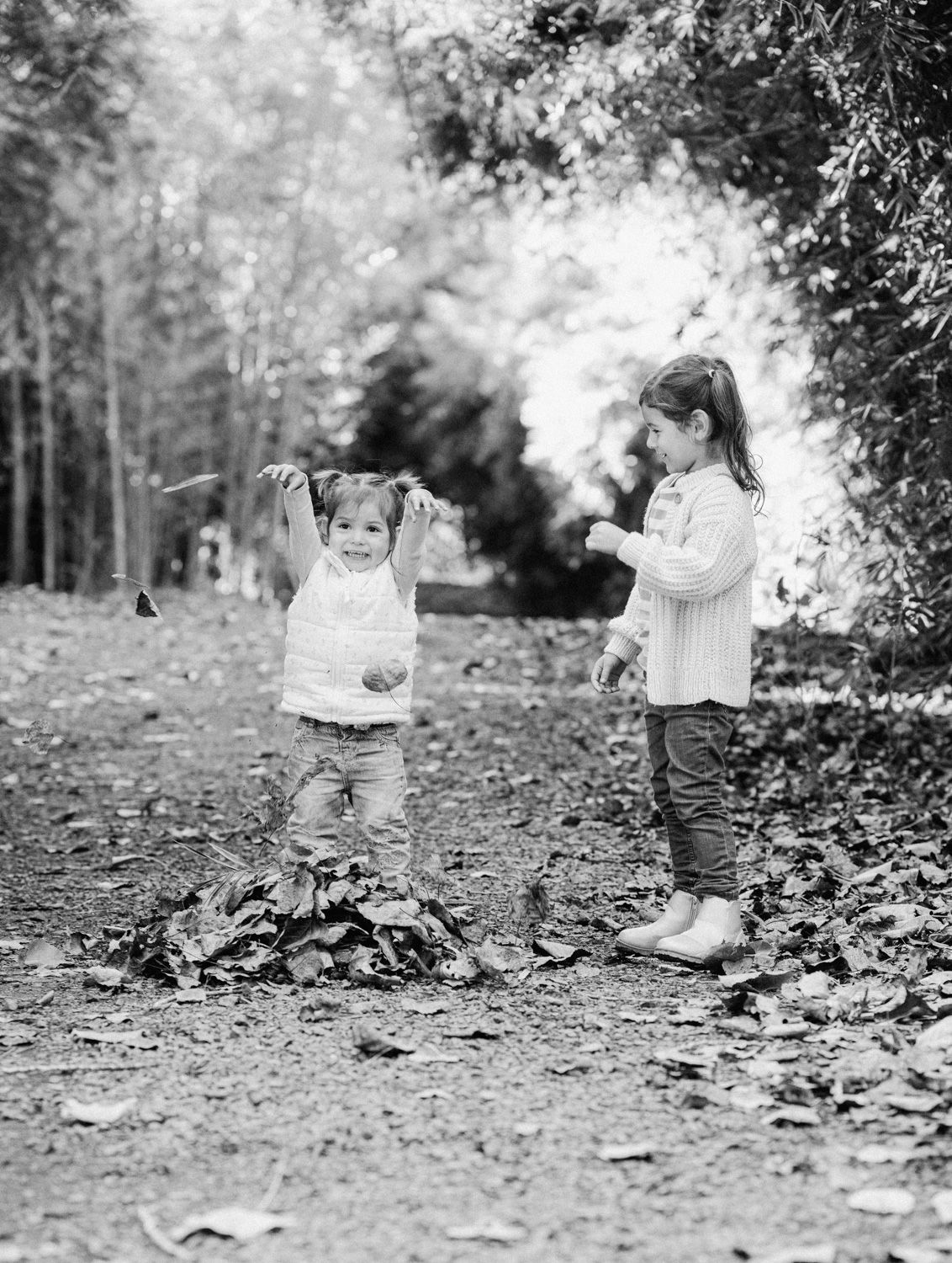 Two young girls playing with autumn leaves on a forest path in a charming black and white photo.