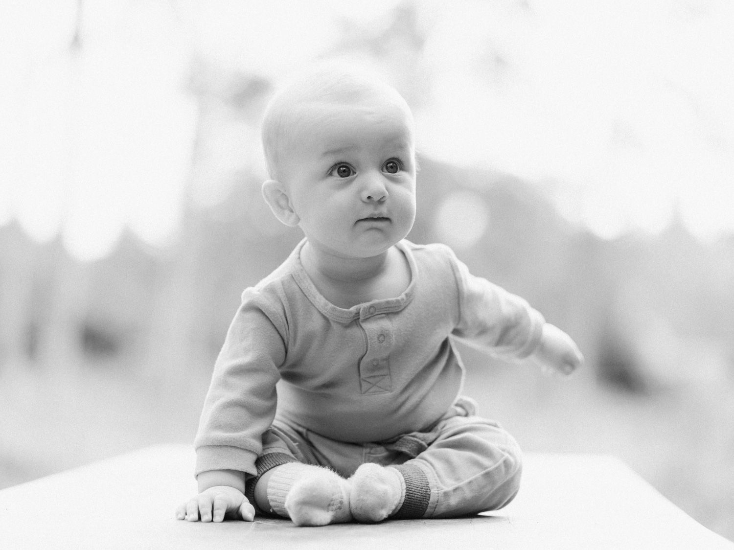 Black and white portrait of an adorable baby sitting cross-legged on a white surface, looking up curiously.