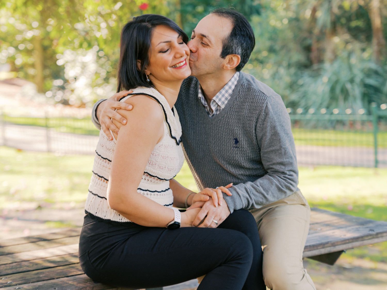 Couple sharing a romantic kiss on a park bench, holding hands outdoors in a sunny green setting.