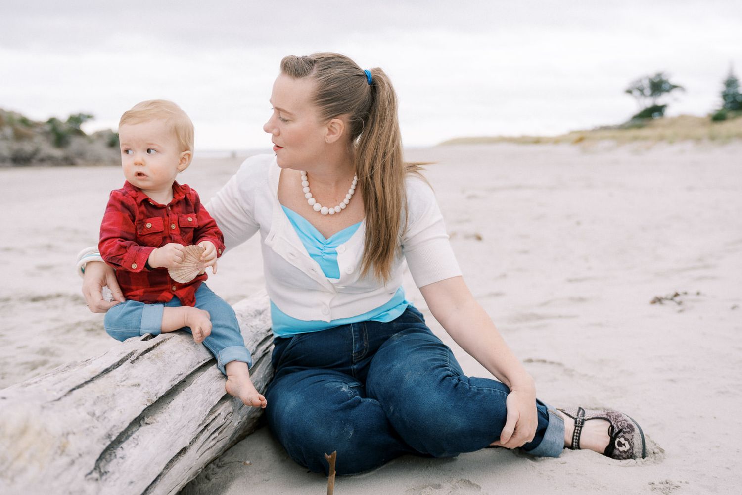 A mother and baby boy sit together on driftwood at a sandy beach, smiling outdoors.