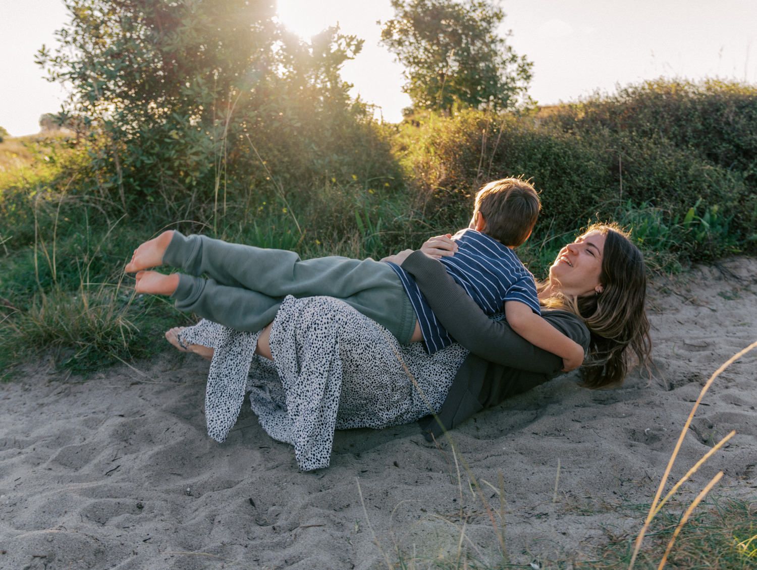 Couple cuddling on sandy beach dunes at golden hour, surrounded by lush green trees and warm sunlight.