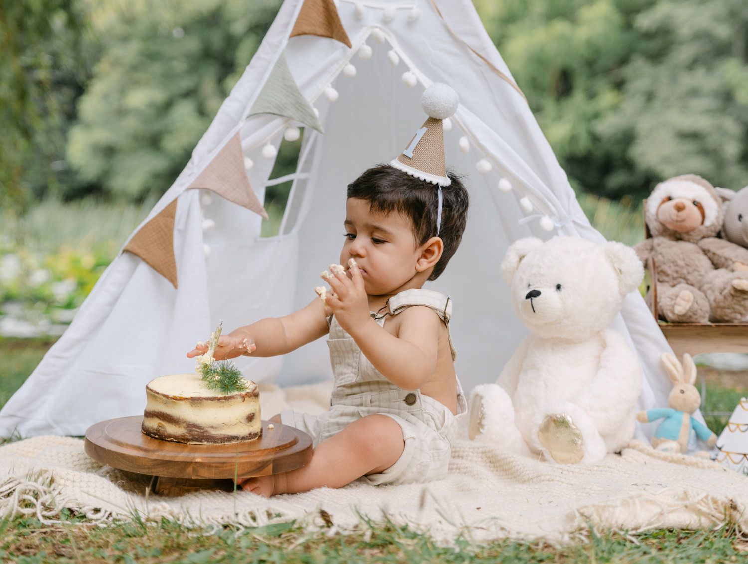 Baby enjoys first birthday cake smash outdoors beside a white teepee with teddy bears on a cozy blanket.