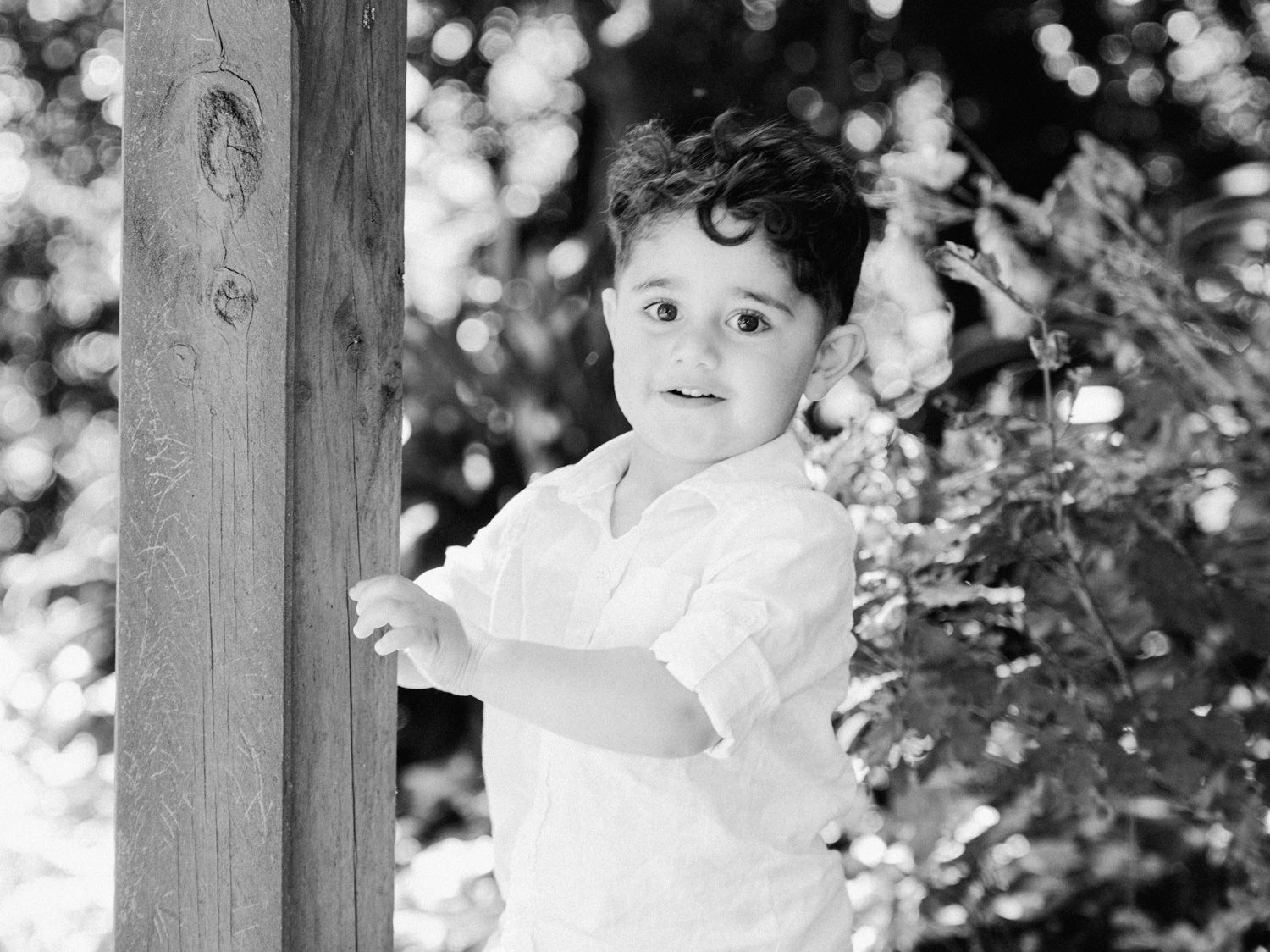 Black and white photo of a young curly-haired boy in a white shirt standing by a wooden post outdoors.