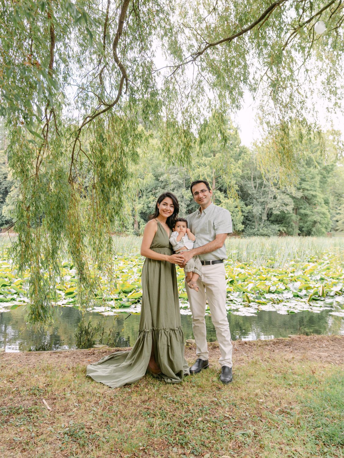 Family of three posing under a weeping willow tree by a lily pad pond, mother in sage green maxi dress holding baby.