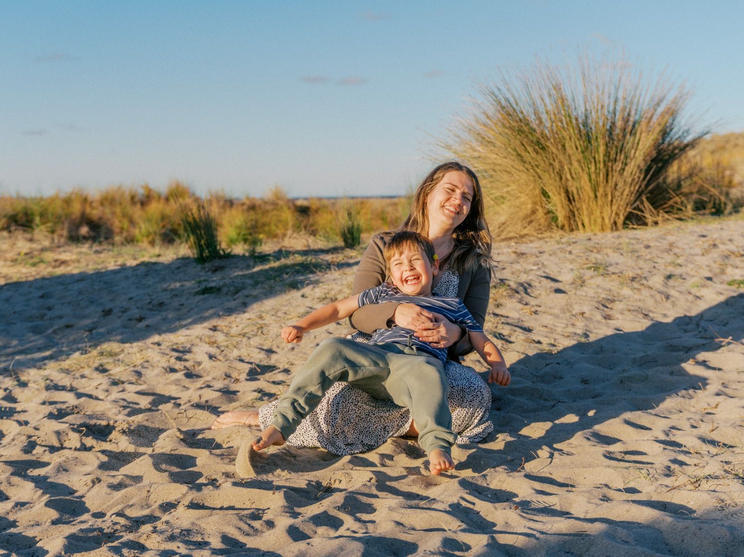 Woman and toddler laughing together on sandy beach dunes at golden hour sunset.