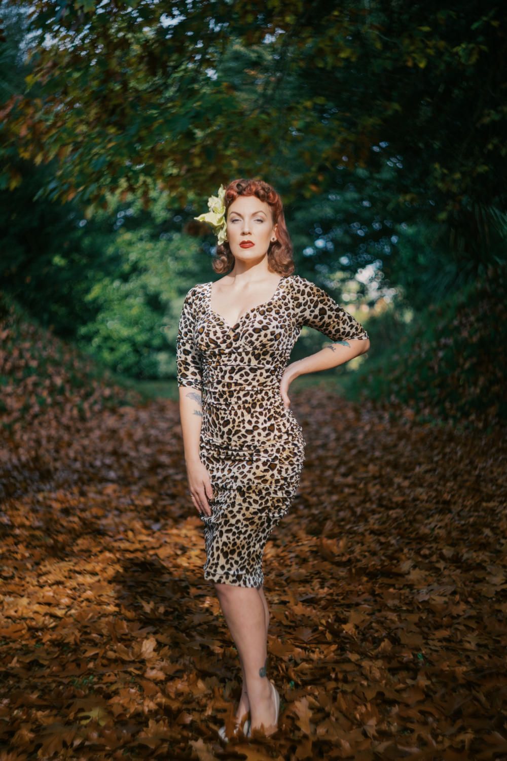 Woman in leopard print dress posing on autumn leaf-covered path surrounded by lush green trees.