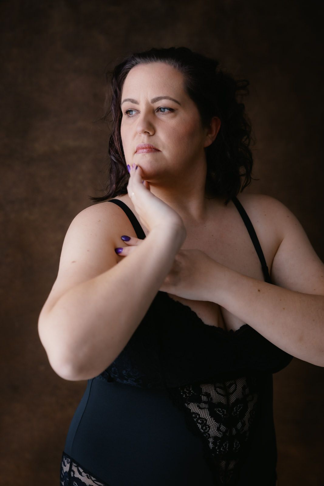 A plus-size woman in black lace lingerie poses thoughtfully against a dark brown studio background.