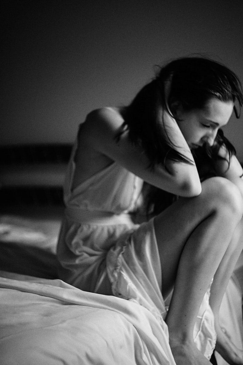 Black and white photo of a woman in white dress sitting on bed, head down, in a pensive, melancholic pose.