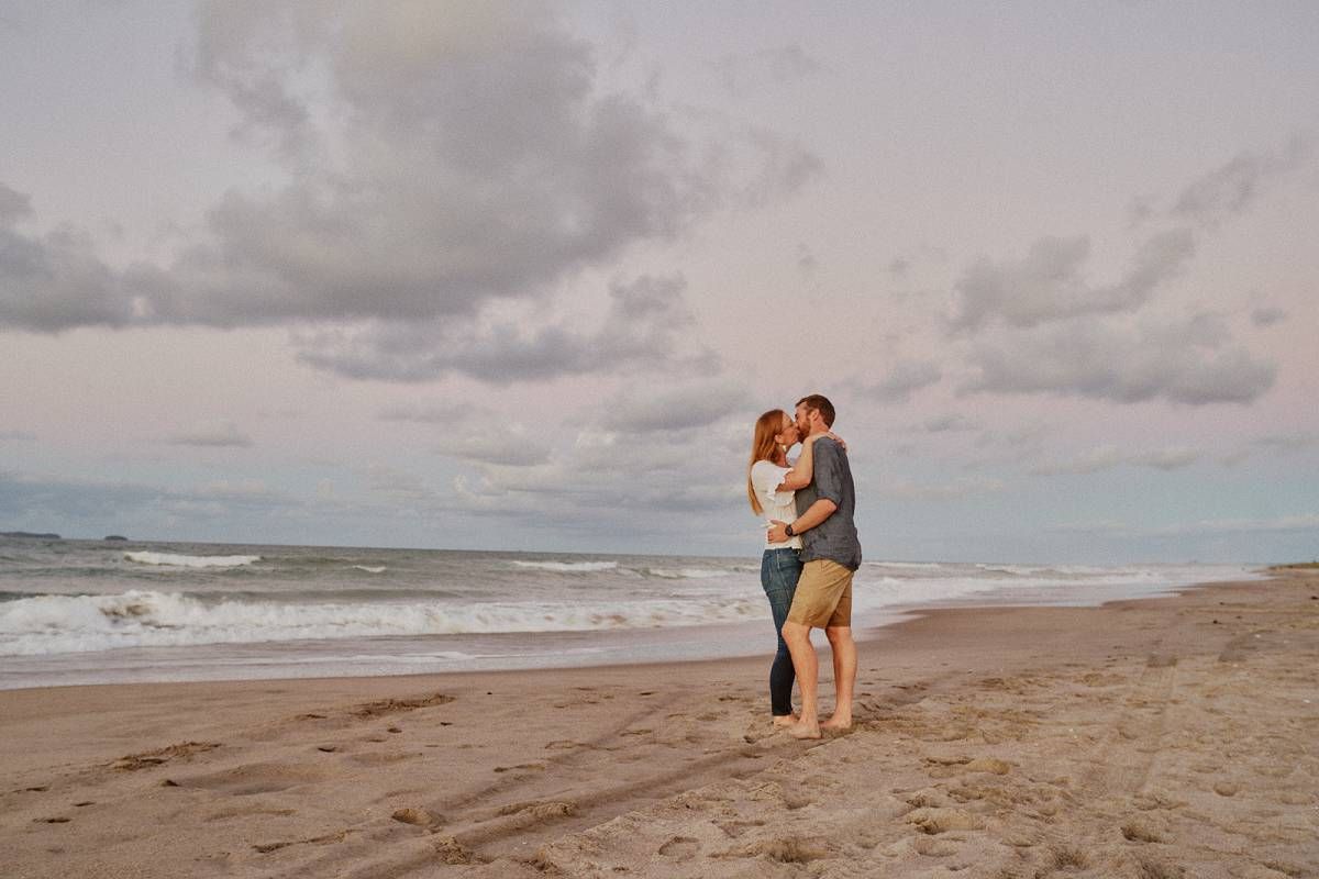 Couple sharing a romantic kiss on a sandy beach at sunset with cloudy skies and ocean waves behind them.