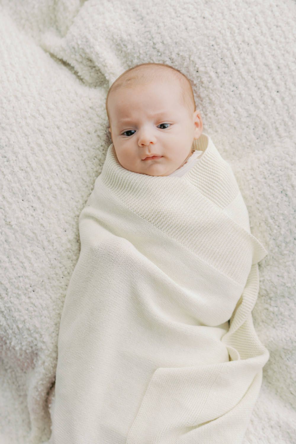 Newborn baby swaddled in a soft cream knit blanket, lying on a white textured surface, looking up calmly.