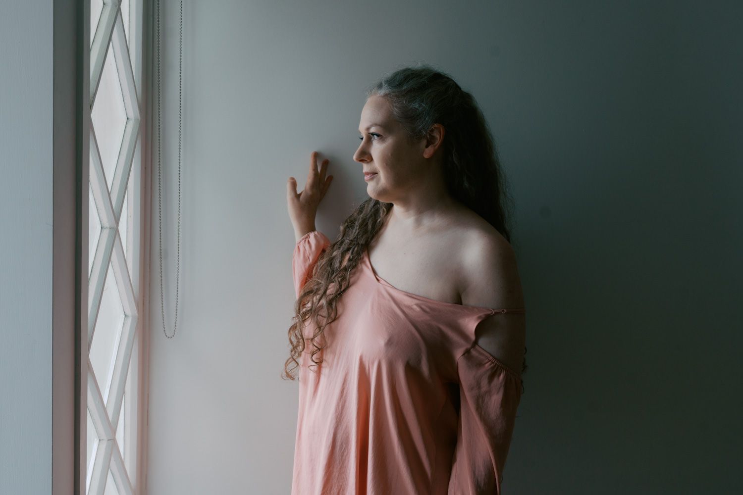 Woman in pink off-shoulder dress gazing thoughtfully out a window with natural light.