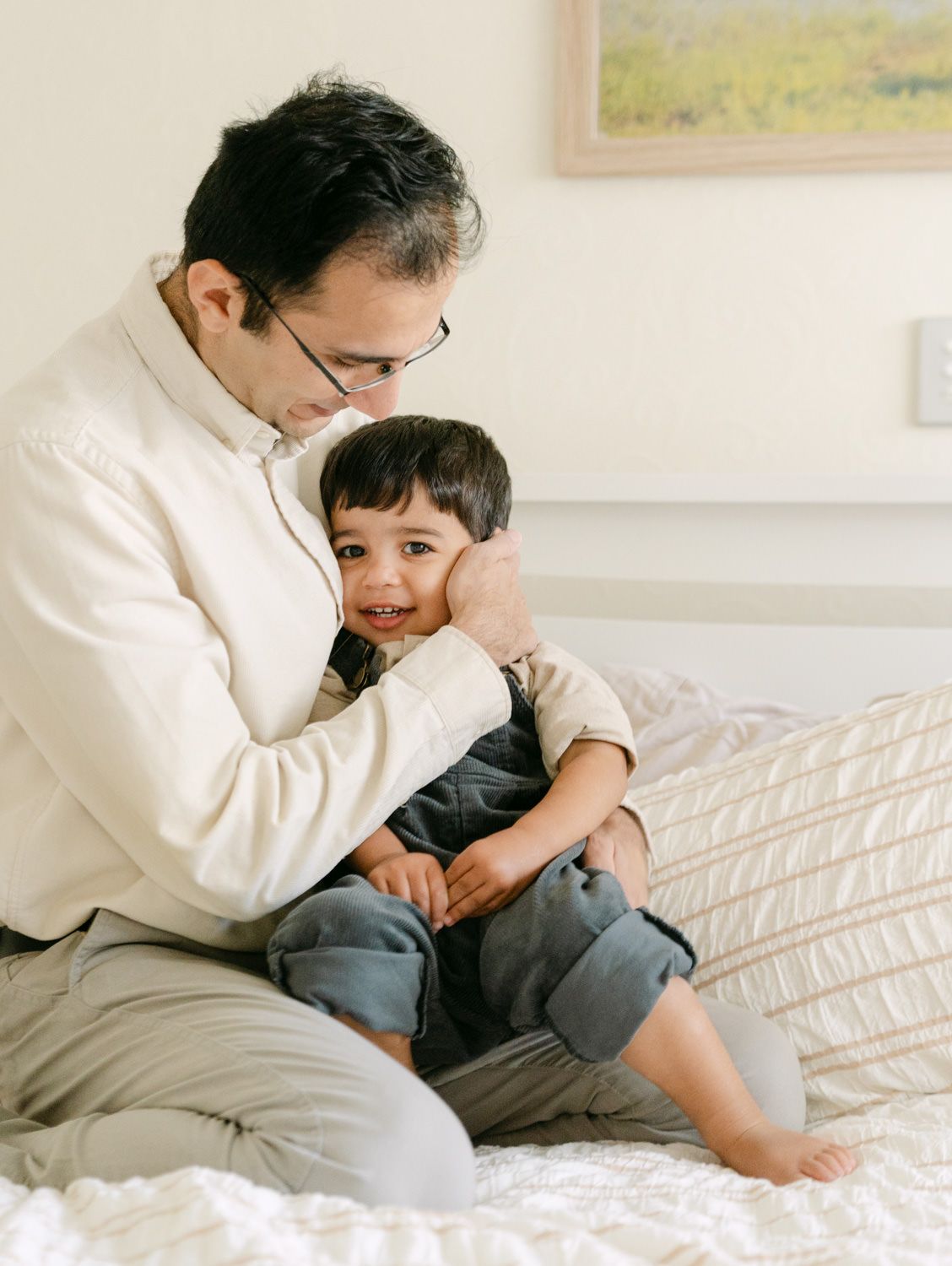 Loving father sitting on bed gently embracing his laughing toddler son in a warm, cozy bedroom.