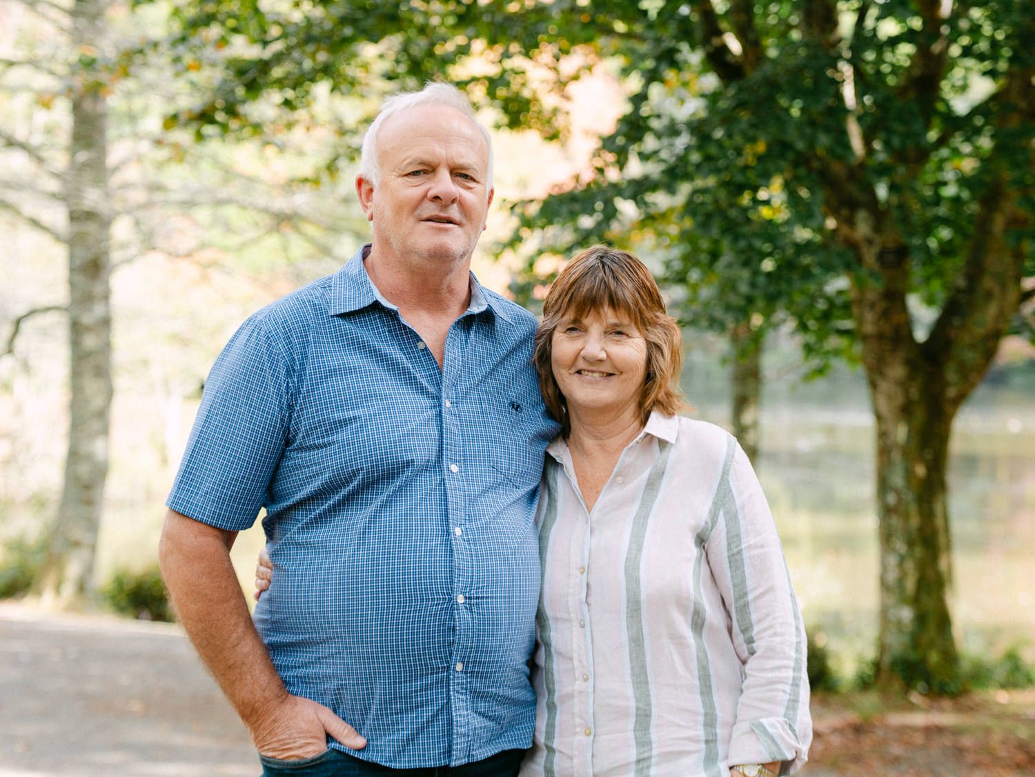 A smiling middle-aged couple standing together outdoors near trees on a sunny day.