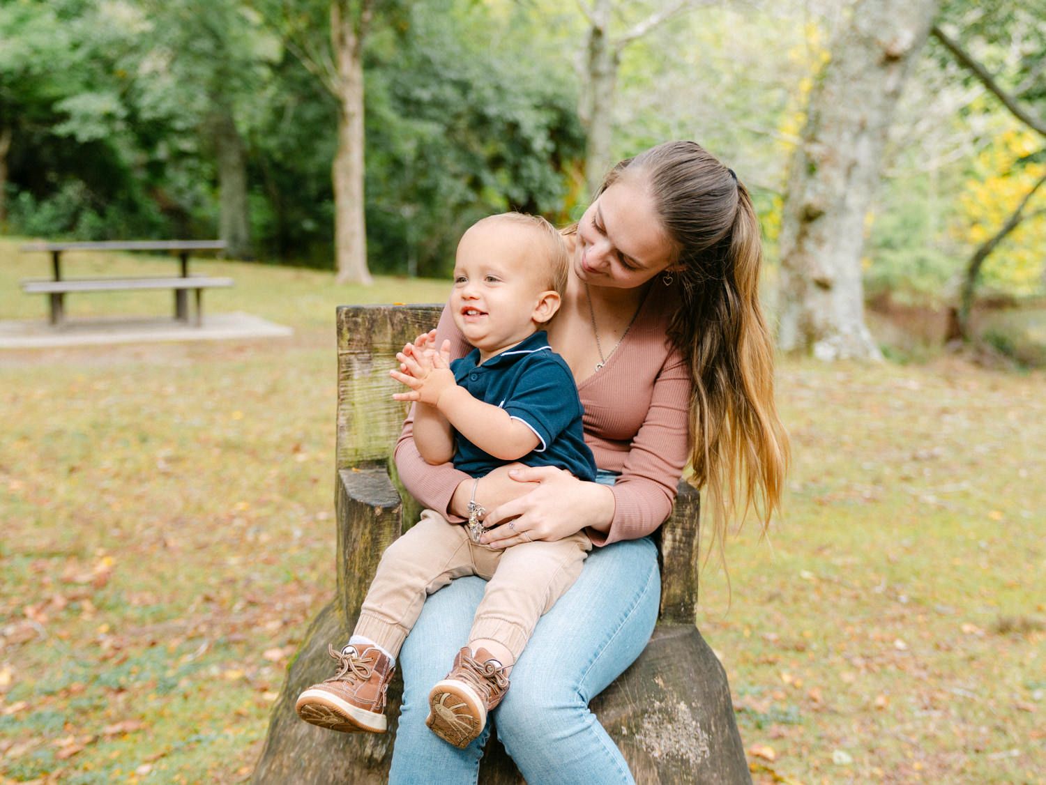 Mother sitting on stone bench in park holding happy toddler baby surrounded by autumn trees.