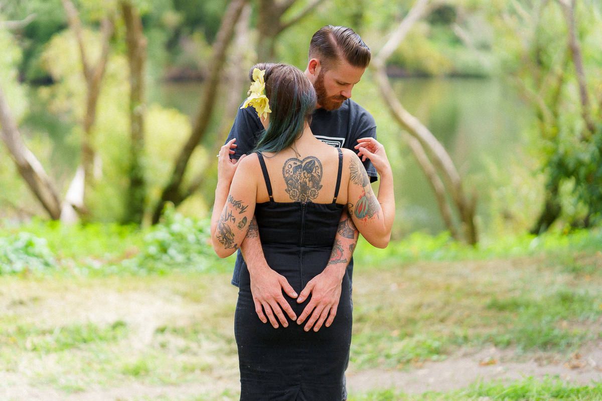 Tattooed couple embracing outdoors near a lake, woman with green hair in black dress, man kissing her head.