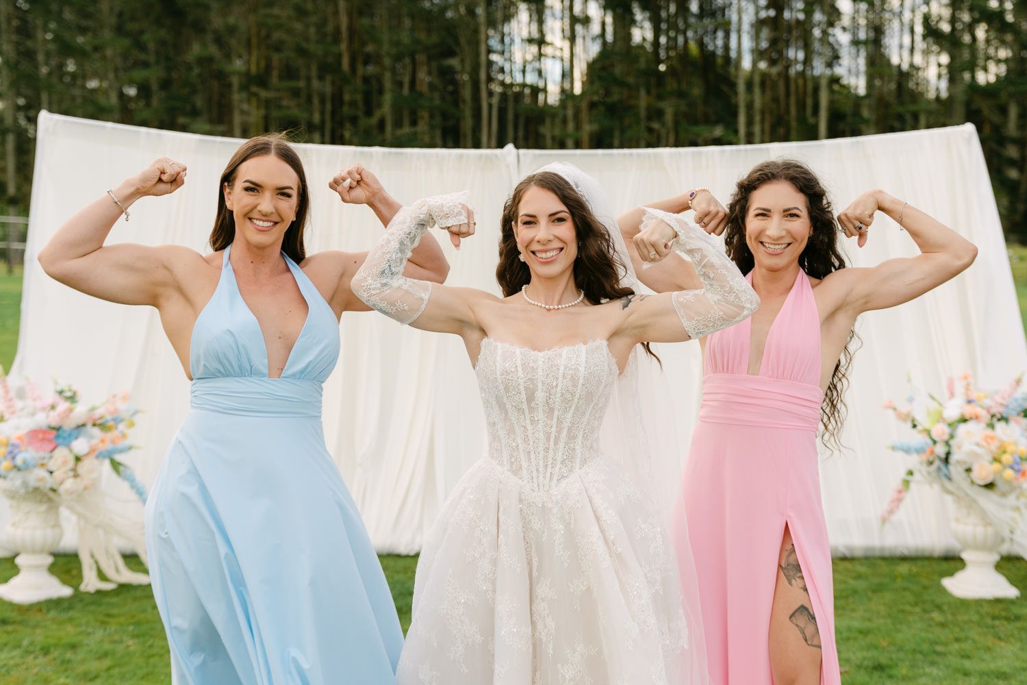Bride in white corset gown flexes biceps with two bridesmaids in blue and pink dresses outdoors.