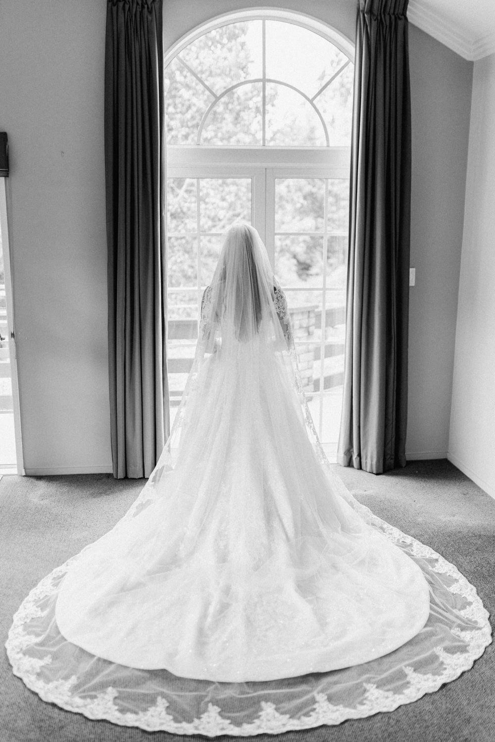 Black and white photo of a bride in a long lace-trimmed gown and veil standing before an arched window.