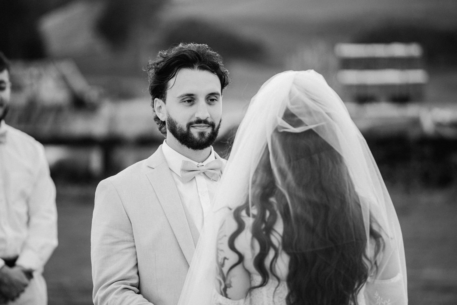 Black and white wedding photo of groom in bow tie smiling at veiled bride during outdoor ceremony.