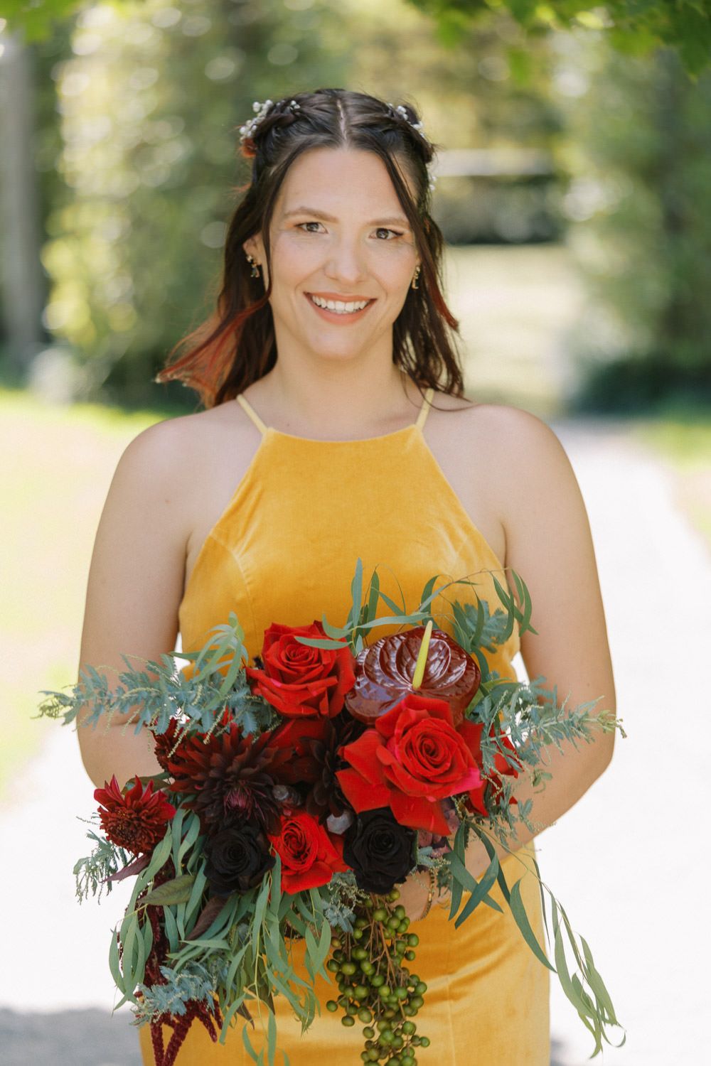 Smiling bridesmaid in yellow velvet dress holding red roses and greenery bouquet outdoors.
