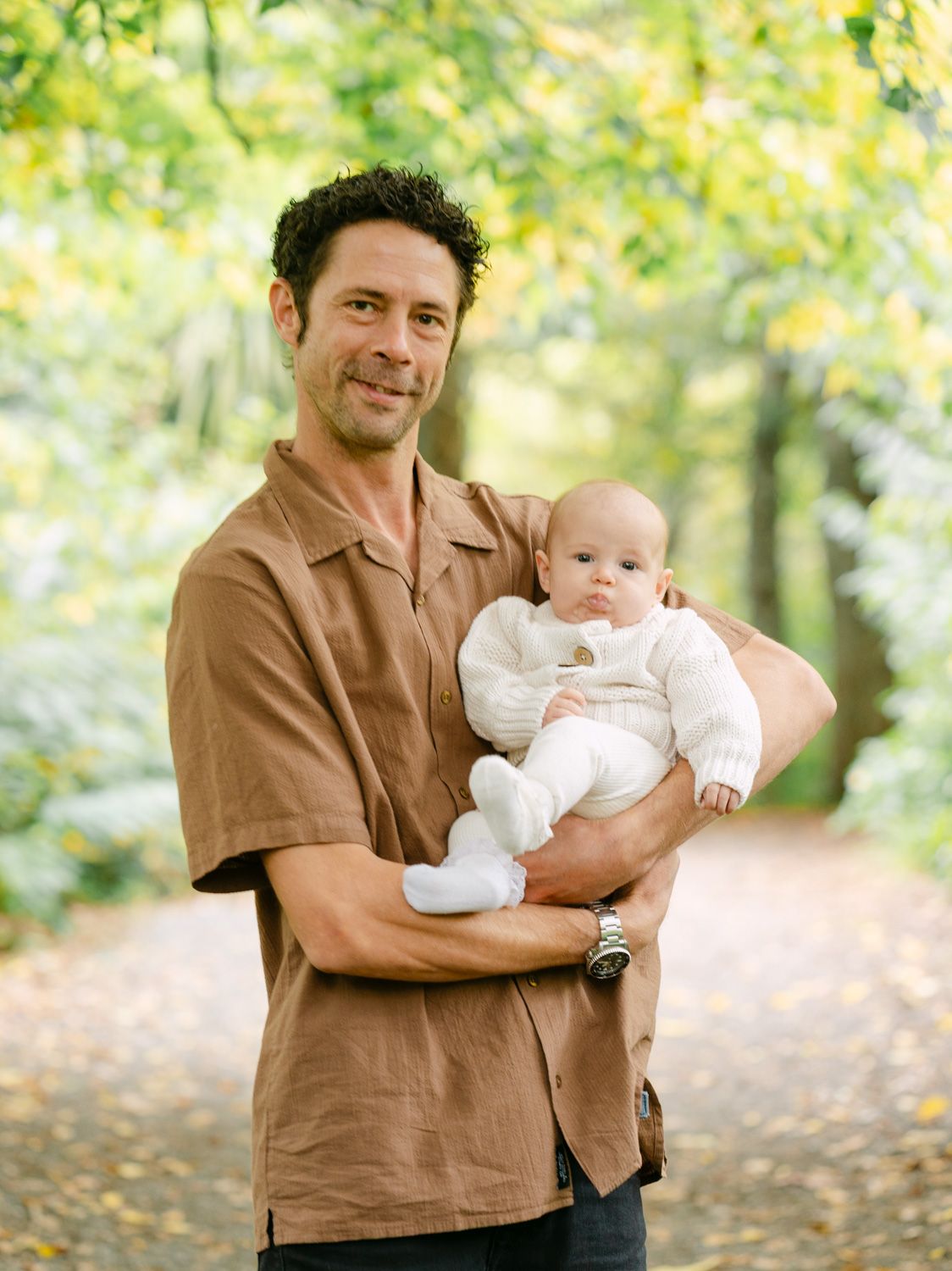 A smiling man in a brown shirt holds a baby in white outdoors on a tree-lined path.