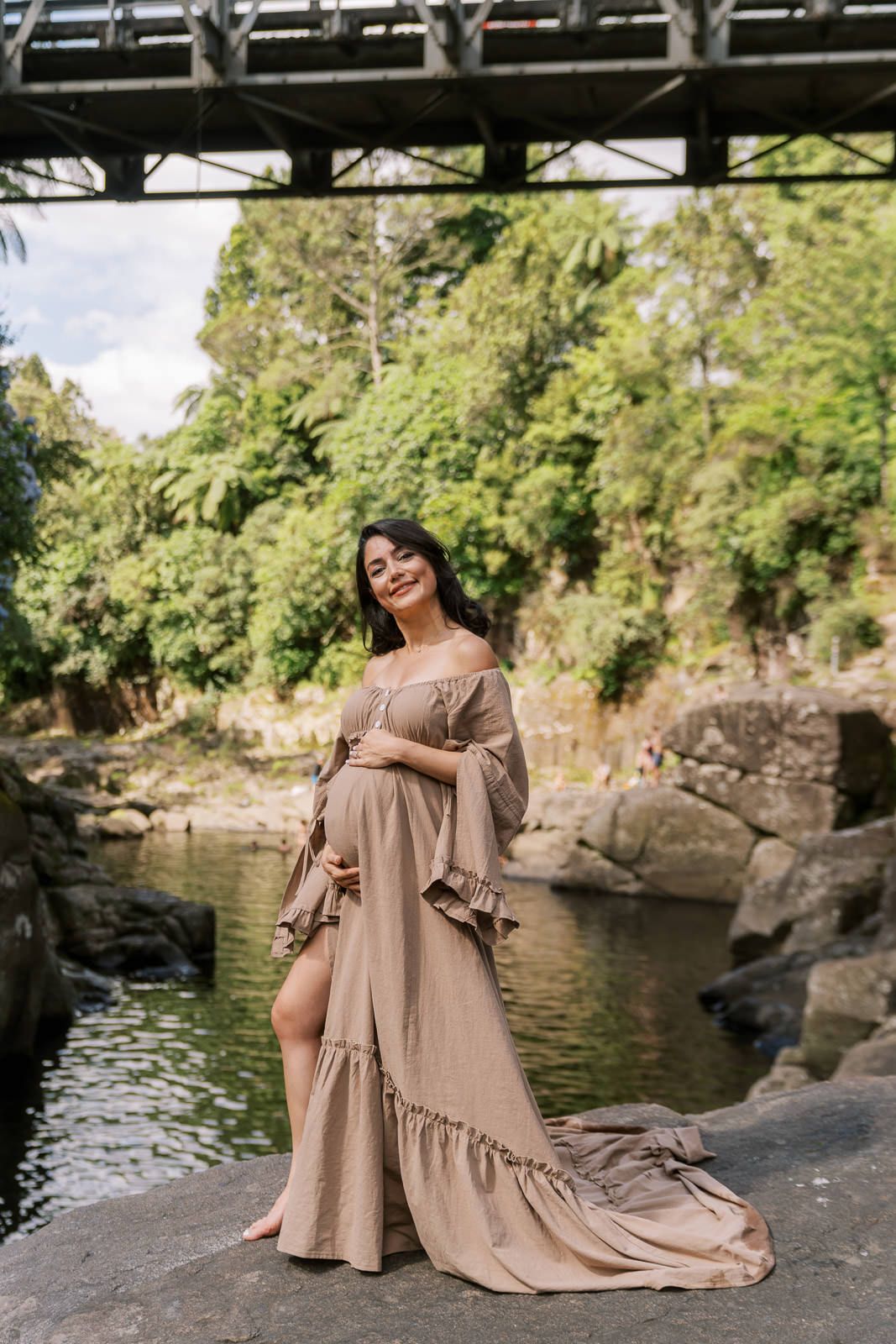 Pregnant woman in elegant beige off-shoulder maxi dress posing on rocks by a serene river surrounded by lush greenery.