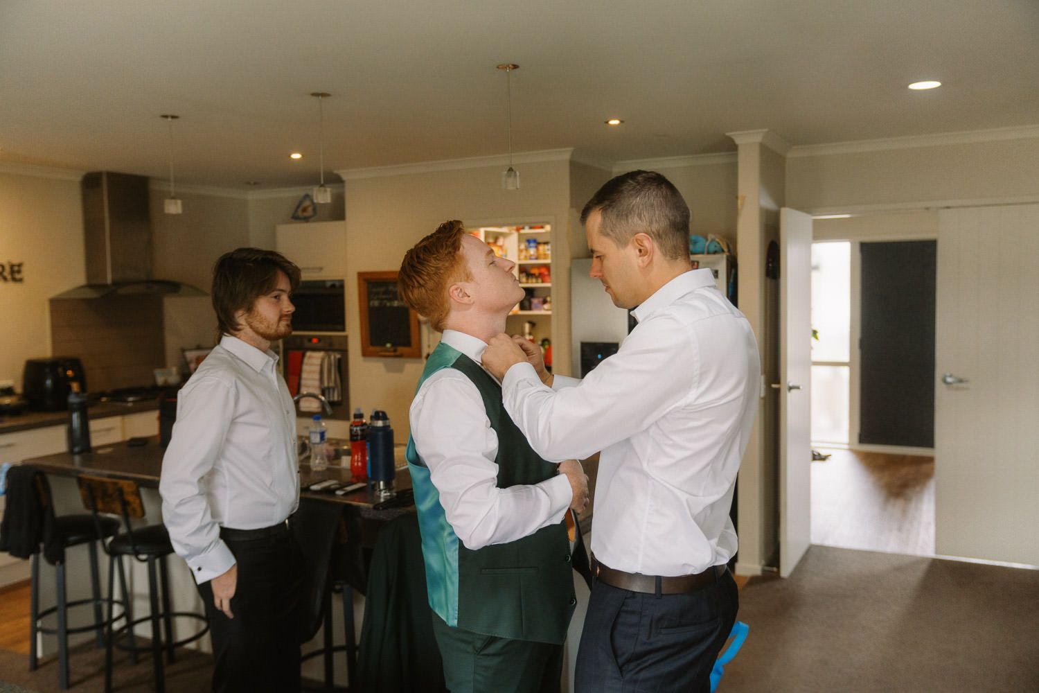 Groom getting his green vest adjusted by groomsman while another watches in a modern kitchen before wedding.