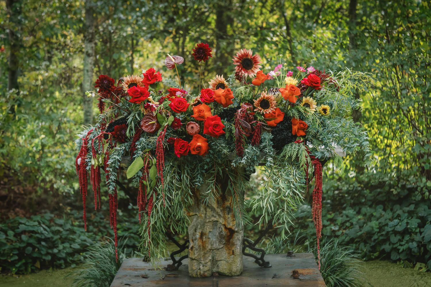 Lush red and orange floral arrangement in a rustic vase displayed outdoors surrounded by green foliage.