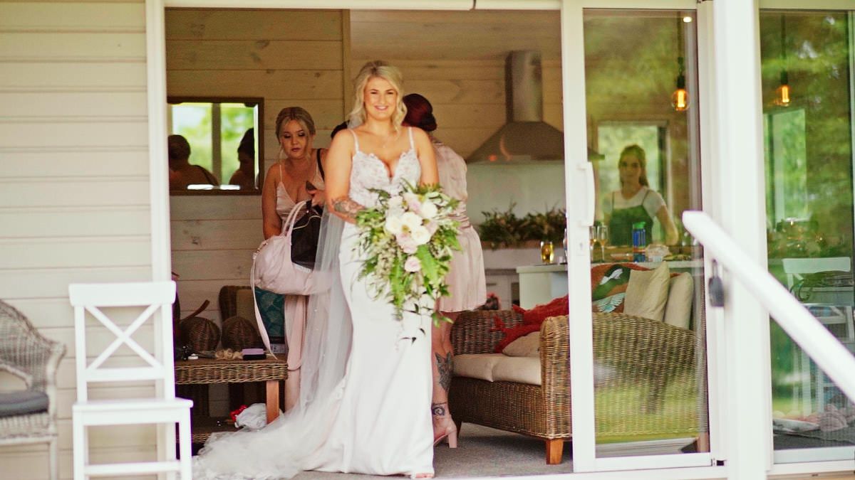 Bride in white gown holding cascading floral bouquet stepping out of a charming cottage on her wedding day.