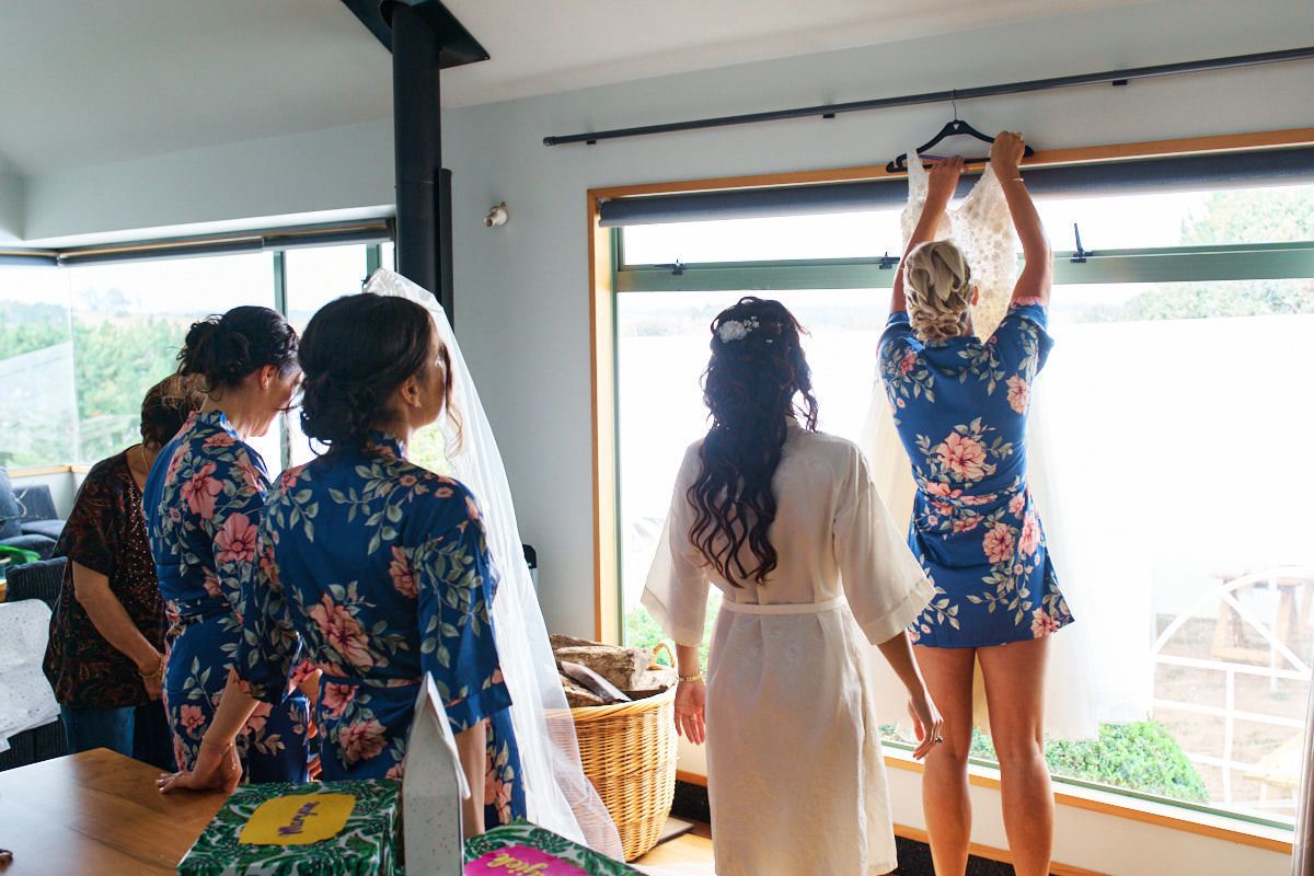 Bridesmaids in blue floral robes gather by window as bride in white robe holds wedding dress on hanger.