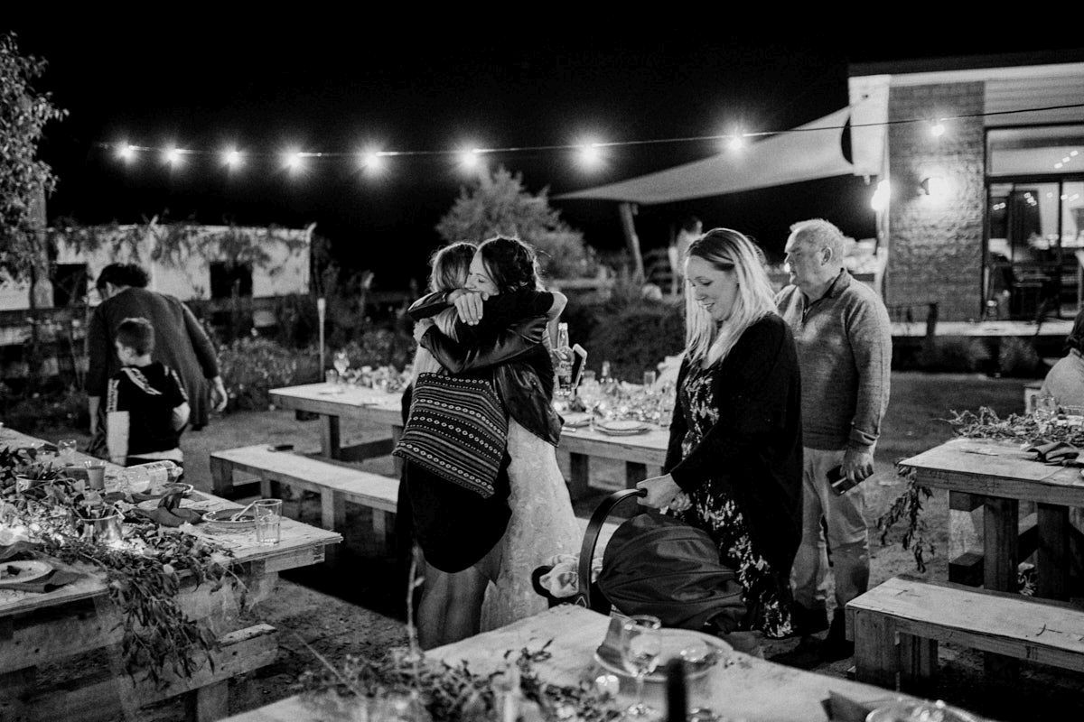 Black and white outdoor evening gathering with string lights, guests hugging and mingling near wooden tables.