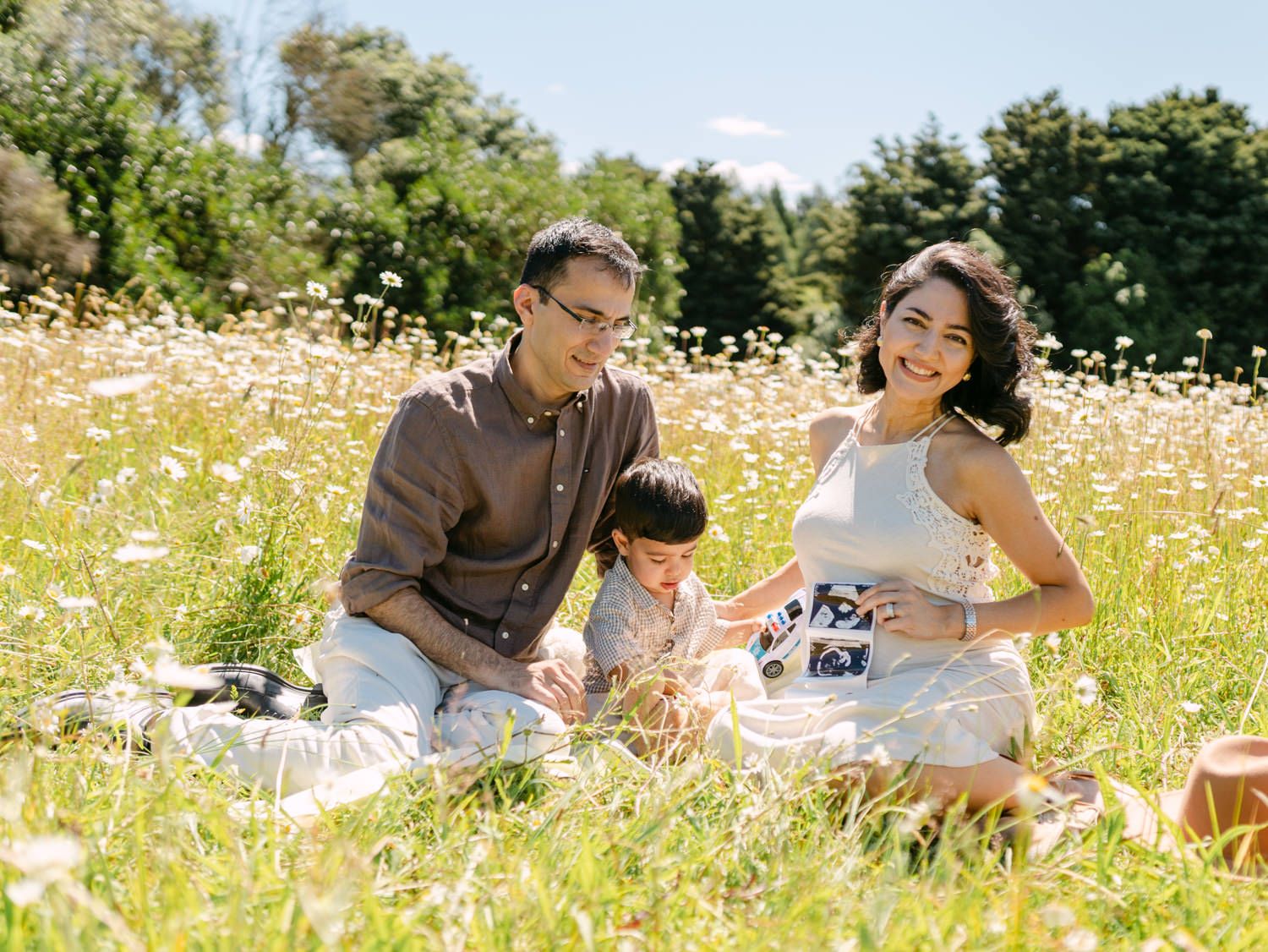 A happy Asian family sitting in a sunny wildflower meadow, with a pregnant mother, father, and young child.