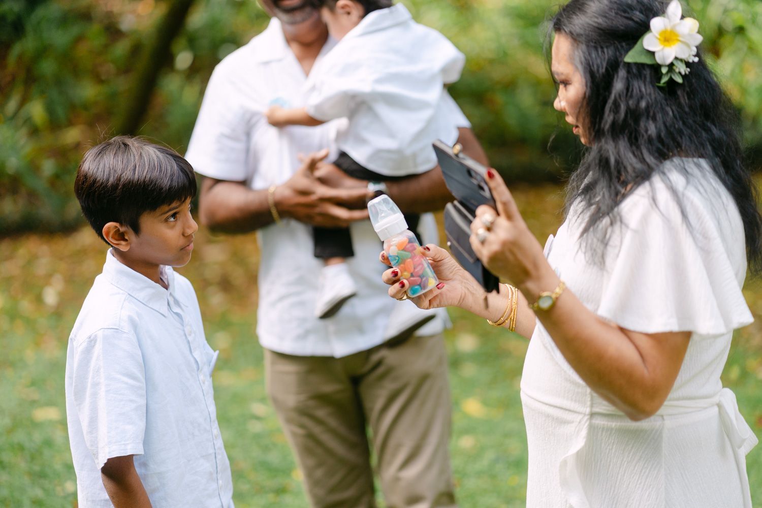 A woman in a white dress shows something to a young boy outdoors during a family gathering in a lush green park.