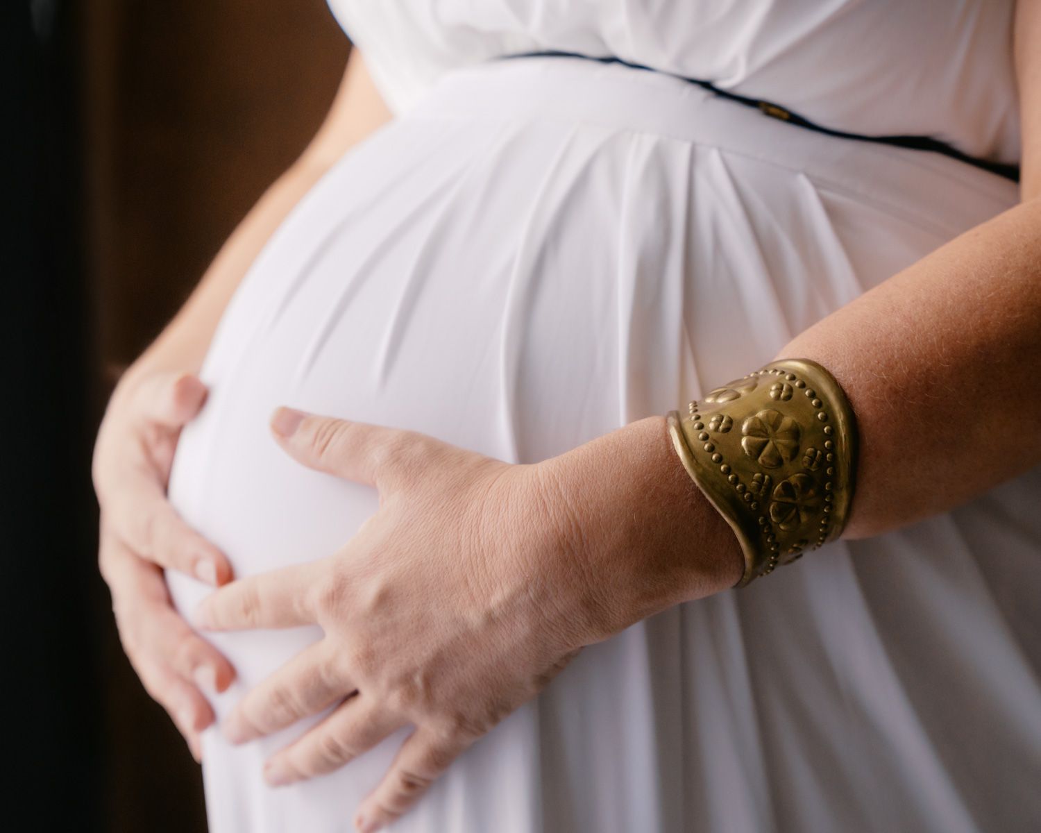 Pregnant woman in white dress cradling baby bump, wearing gold cuff bracelet, close-up maternity photo.